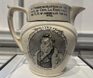 An off-white creamer pitcher with a black and white drawn image of the Marquis de Lafayette. The inscription reads "In Commemoration of the Visit of Gen. LaFayette to the U.S. of Ameria in the Year 1824."