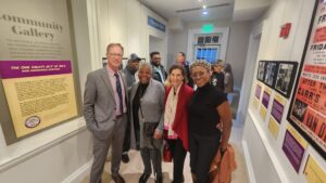 Four people stand in a line in a museum exhibit hall. The walls are lined with photos and descriptive text panels.